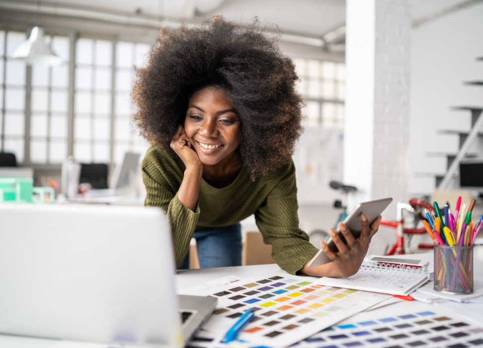 Woman at table holding mobile device while looking at laptop.