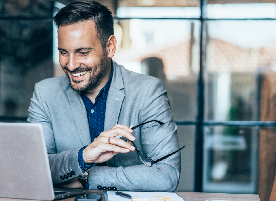 Man sitting at desk holding glasses looking at laptop.