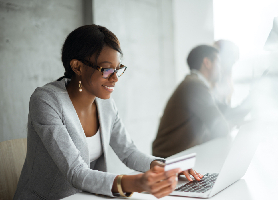 Woman sitting at table wearing glasses, looking at laptop while holding credit card.