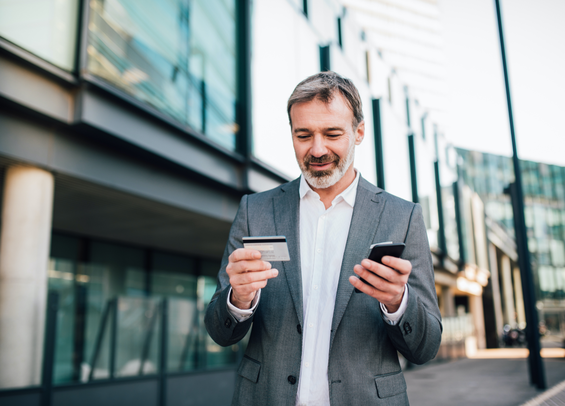 Man standing outside of building looking at debit card and mobile device.