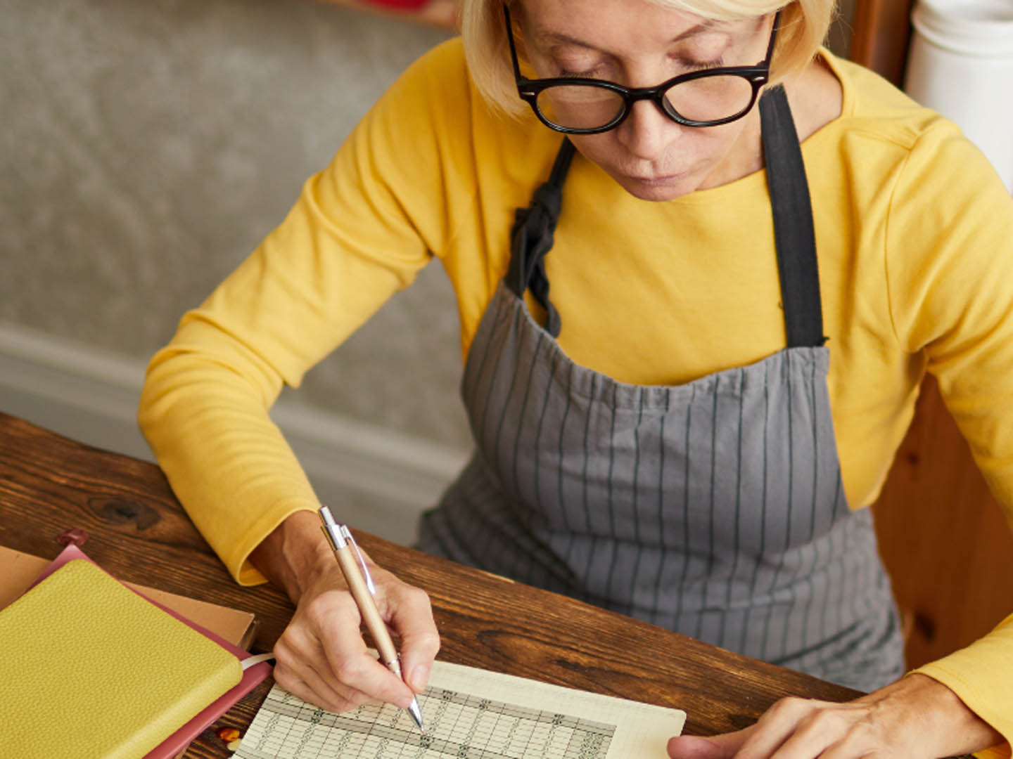 Woman wearing glasses and apron sitting at table writing in ledger