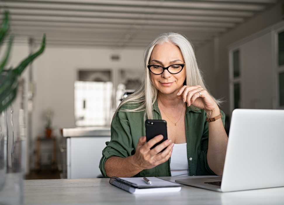 Mature woman sitting at desk with laptop looking at mobile phone