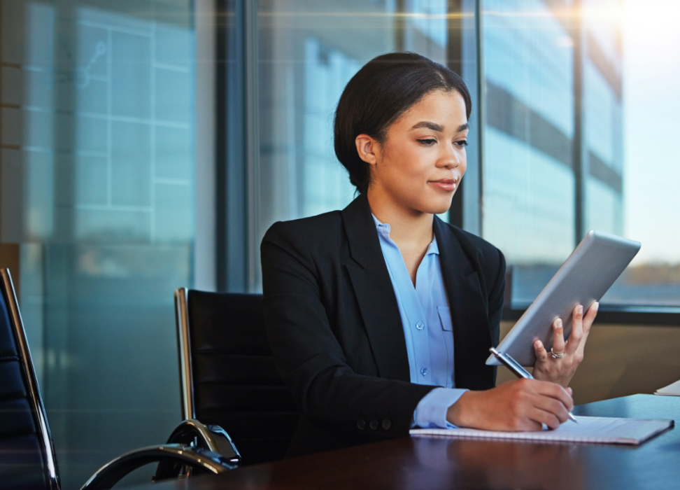 Woman sitting at table taking notes while looking at tablet.