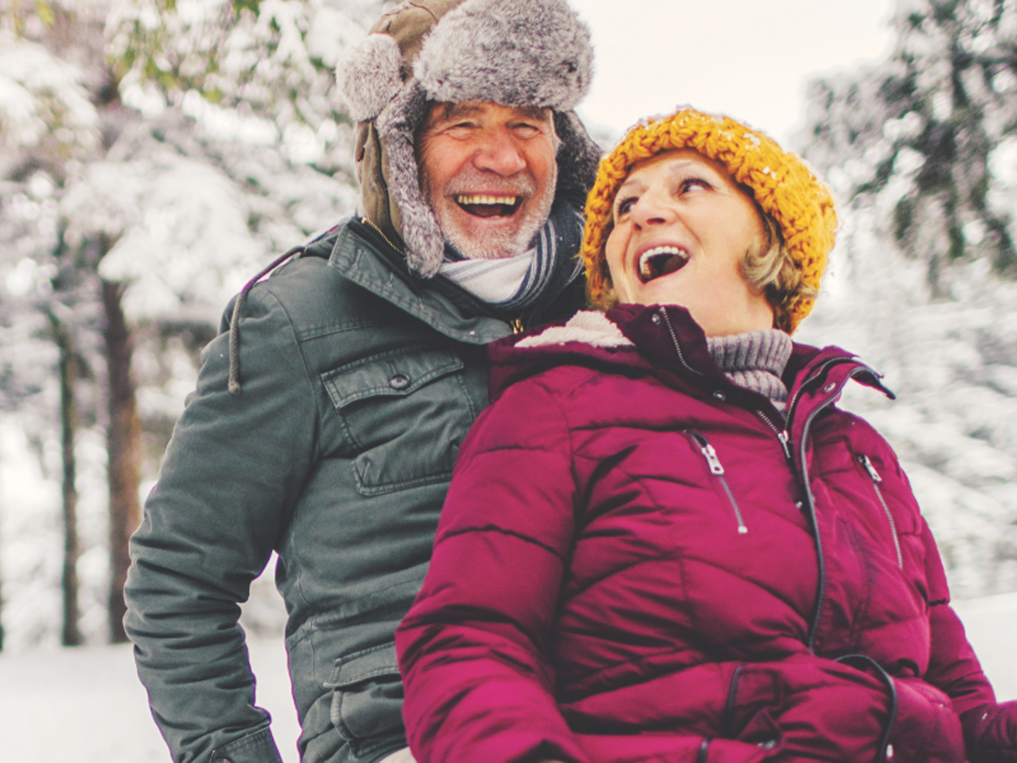 Man and woman laughing wearing winter hat and jackets while sitting in the snow.