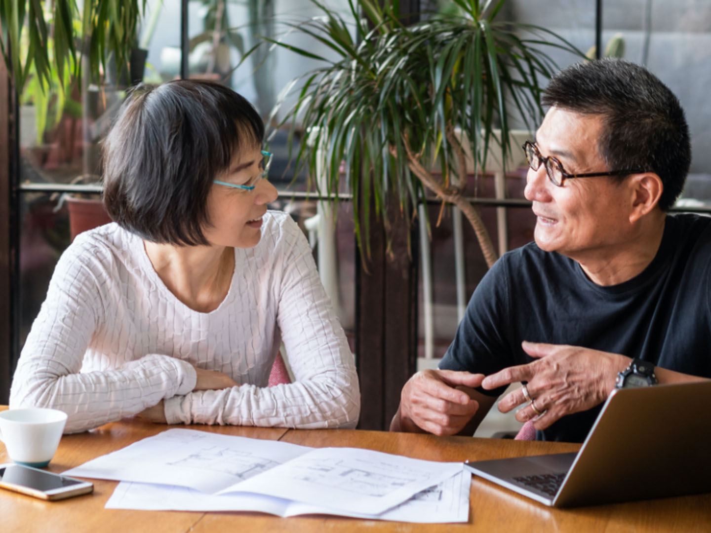 Man and woman wearing glasses sit at table with laptop and paper.