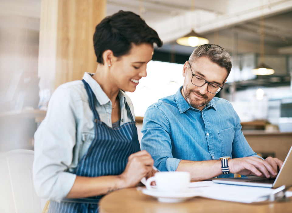 Woman and man sitting at table in cafe with laptop looking at notepad.