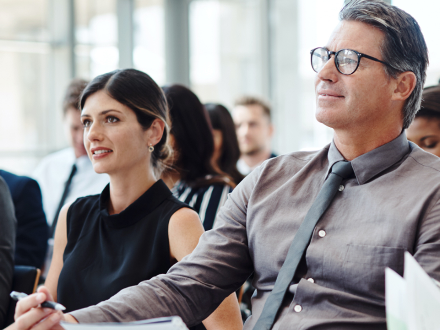 Man wearing glasses and woman wearing sleeveless shirt sitting taking notes at a group meeting.